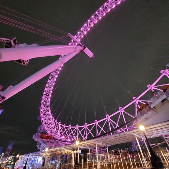 Ein Riesenrad mit einer lila Lichtshow ist nachts zu sehen, wobei seine Kabinen am Rad befestigt sind. Das Rad wird von mehreren Seilen gestützt.