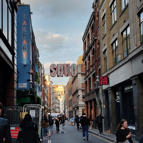 Eine belebte Straße mit einem 'Soho'-Schild über der Straße. Menschen gehen vorbei und Geschäfte befinden sich auf beiden Seiten. Eines hat ein Neon-Schild 'Be at One Cocktail Bar'.
