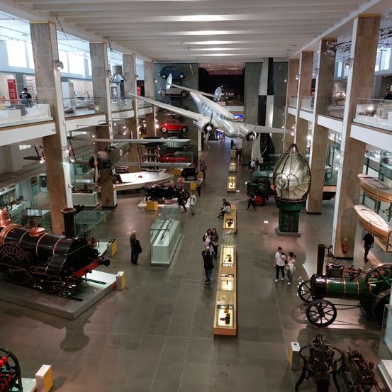 Ein Luftbild einer Innenausstellung mit zahlreichen alten Flugzeugen, Autos und Zügen. Besucher gehen durch den geräumigen Saal und betrachten die historischen Exponate.