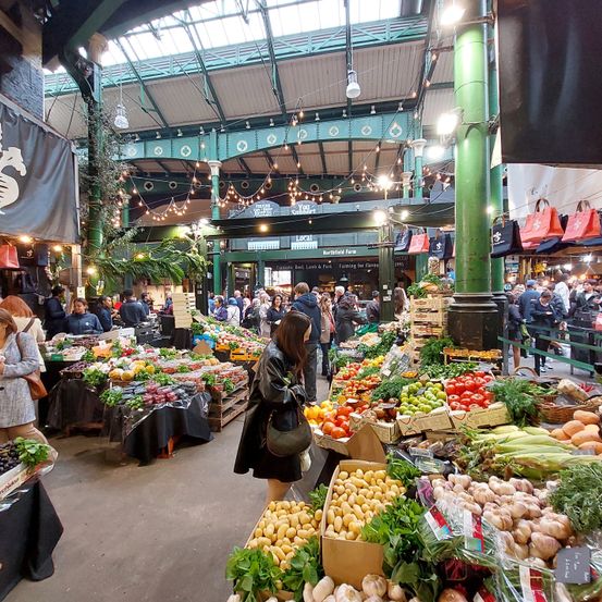 Ein geschäftiger Innenmarkt voller frischem Obst, Gemüse und Kräutern. Menschen kaufen ein, und verschiedene Stände präsentieren ihre Waren. Die Decke ist mit hängenden Lichtern und dekorativen Elementen geschmückt.