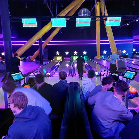 Eine Bowlingbahn mit lila Beleuchtung, in der Menschen spielen und zusehen. Bowling-Pins und Bälle sind vorhanden, zusammen mit Monitoren, die die Punkte anzeigen.