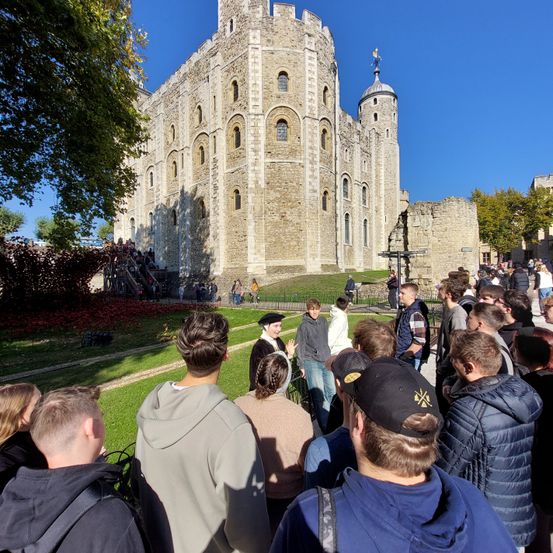 Eine Gruppe von Menschen versammelt sich vor einer historischen Burg und lauscht einem Führer. Die Burg hat einen Turm und ist von Bäumen und Gras umgeben.
