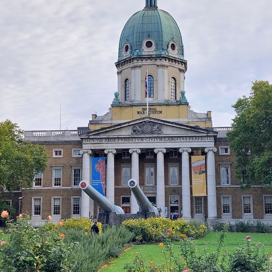 Das Imperial War Museum in London mit einer grünen Kuppel und zwei Kanonen davor, mit Bannern und üppiger Begrünung.