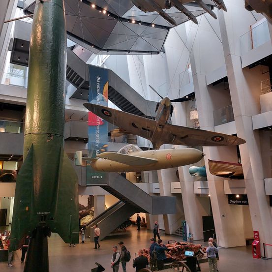 Eine Ausstellungshalle mit Flugzeugen und Raketen, die von der Decke hängen. Banner hängen in der Mitte. Leute gehen herum.