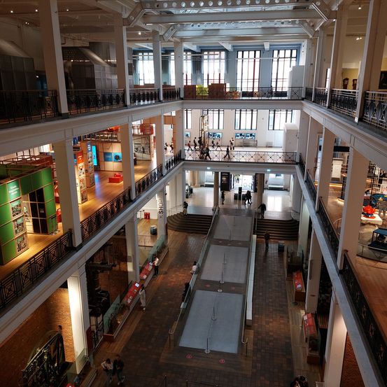 Ein mehrstöckiger Museumsinnenbereich mit Besuchern, die verschiedene Ausstellungen erkunden. Der zentrale Gang wird von Treppen flankiert, und die oberen Ebenen bieten eine Vielzahl von Ausstellungen und Galerien.