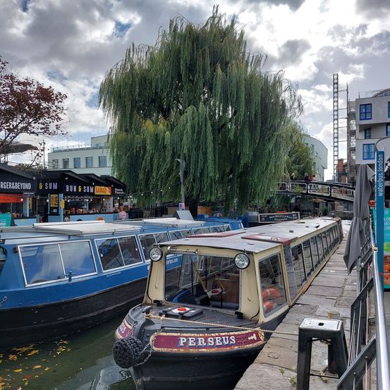 Ein Kanal mit angedockten Booten, eines davon Perseus, unter einem bewölkten Himmel mit Gebäuden und einer Brücke im Hintergrund.