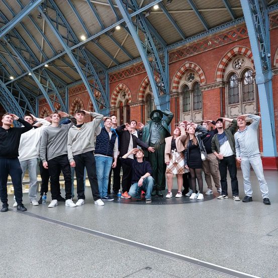Eine Gruppe von Menschen posiert für ein Foto in einer Halle mit einer Statue in der Mitte. Sie alle lächeln und scheinen sich zu amüsieren.