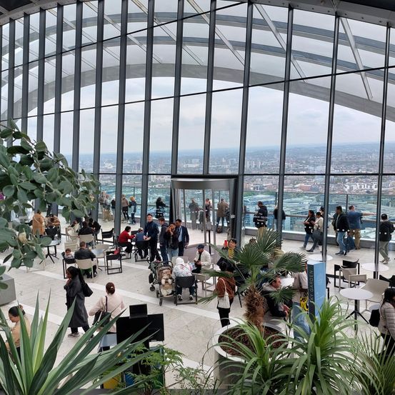 Ein Indoor-Gartencafé mit Panoramablick auf die Stadt. Viele Menschen sitzen an Tischen und gehen herum, einige machen Fotos der Stadtlandschaft durch große Fenster.