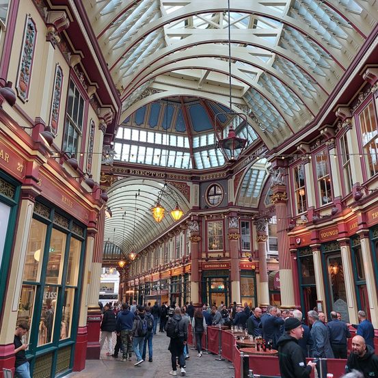 Eine geschäftige überdachte Einkaufspassage mit hohen Decken, Glasdächern und mehreren Geschäften. Menschen gehen und stehen herum, und einige sitzen an Außencafés.