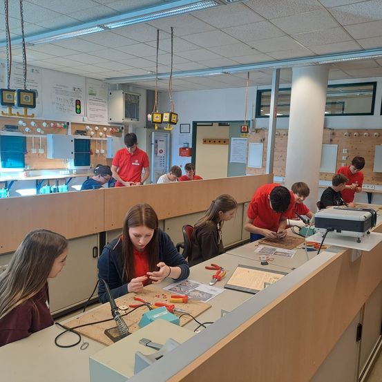 Schüler arbeiten in einem Klassenzimmer mit einem Holztisch und verschiedenen Werkzeugen an Lötprojekten. Einige tragen rote Hemden, einer hat einen Rucksack. Lichter hängen von der Decke.