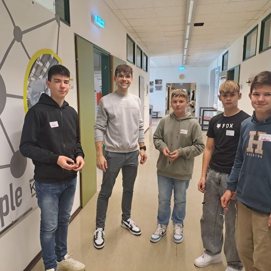 Fünf Jungen stehen in einem Flur. Sie tragen alle Freizeitkleidung und Sneakers. Sie scheinen in ein Gespräch vertieft zu sein.