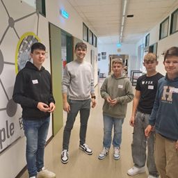 Fünf Jungen stehen in einem Flur. Sie tragen alle Freizeitkleidung und Sneakers. Sie scheinen in ein Gespräch vertieft zu sein.