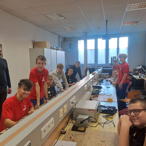 Eine Gruppe junger Schüler in roten Hemden arbeitet in einem Klassenzimmer an einem Projekt mit verschiedenen elektronischen Geräten.