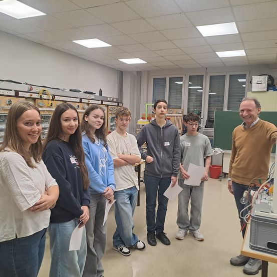 Eine Gruppe von Schülern und einem Lehrer in einem Labor, sie lächeln und posieren für ein Foto mit Papieren in der Hand.