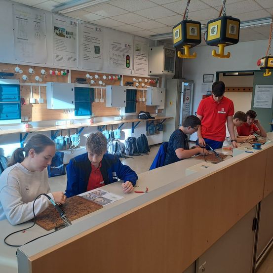 Eine Gruppe von Schülern arbeitet in einem Klassenzimmer an einem Lötprojekt mit verschiedenen elektrischen Komponenten und Werkzeugen.