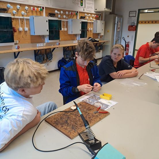 Vier Jungen sind in einer Werkstatt und beschäftigen sich mit Löten. Sie sitzen um einen Tisch herum, vor ihnen liegen Werkzeuge und Materialien.