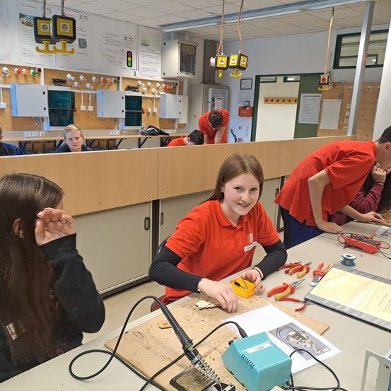Mehrere Schüler arbeiten in einem Labor an Elektronik und tragen rote Hemden. Eine Frau lächelt und schaut auf einen Lötkolben. Verschiedene Werkzeuge und Papiere liegen auf dem Tisch.