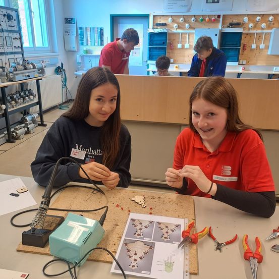 Zwei Mädchen sitzen an einem Tisch in einer Werkstatt, eine benutzt ein Löteisen für ein Projekt, die andere hält ein kleines elektronisches Bauteil. Verschiedene Werkzeuge sind um sie herum platziert. Im Hintergrund steht eine Person in einem roten Shirt an einer Theke.