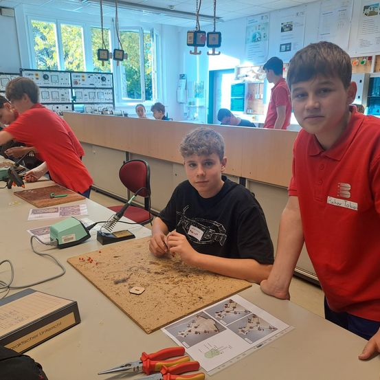 Mehrere Schüler befinden sich in einem Klassenzimmer und nehmen an einem Workshop teil. Zwei Jungen sind prominent zu sehen, einer sitzt und lötet, der andere steht neben ihm. Werkzeuge und Materialien liegen auf dem Tisch vor ihnen.