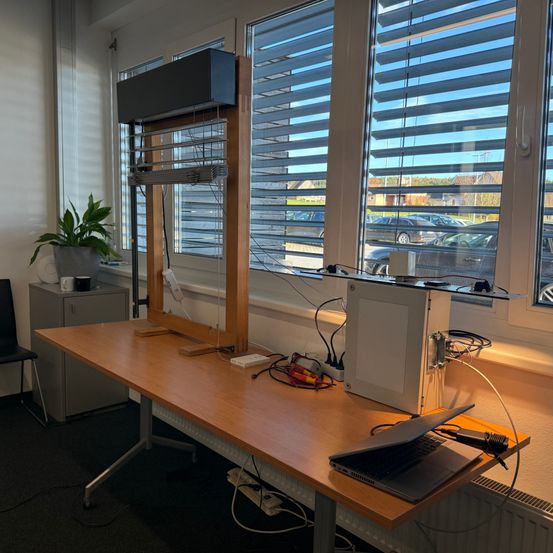 Ein Büroaufbau mit Schreibtisch, Laptop und verschiedenen elektronischen Geräten. Eine Pflanze steht auf einem kleinen Schrank neben einem Fenster mit Jalousien. Draußen sind mehrere Autos geparkt.