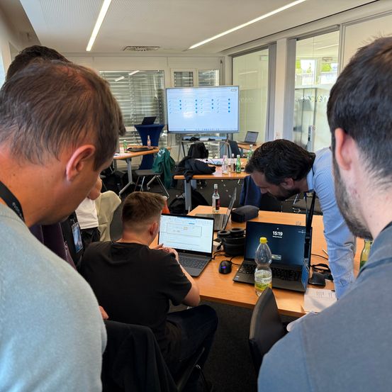 In einem Raum sind mehrere Personen um einen Schreibtisch mit Laptops versammelt, ein Mann in einem schwarzen T-Shirt sitzt, während ein anderer Mann in einem blauen Hemd steht und einen Monitor betrachtet.