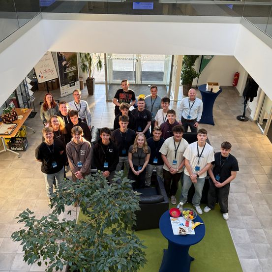 Eine Gruppe von Menschen posiert für ein Foto in einer modernen Lobby, umgeben von Pflanzen und einem Tisch mit Obst. Einige Personen tragen Namensschilder.