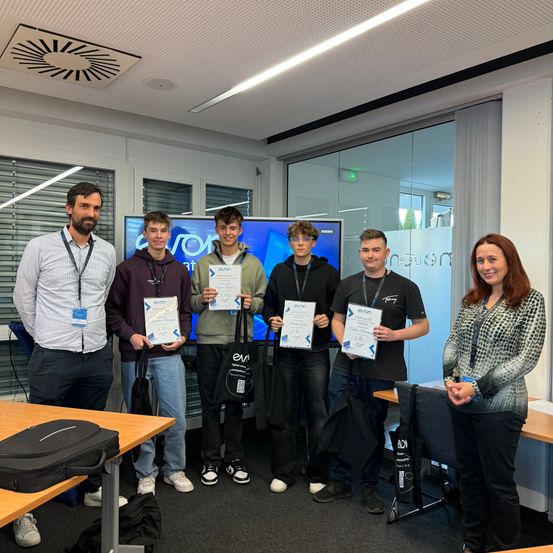 Fünf junge Männer und eine Frau stehen in einem Raum und halten Urkunden in der Hand. Sie lächeln und scheinen eine Errungenschaft zu feiern. Hinter ihnen befindet sich ein Monitor mit Text.
