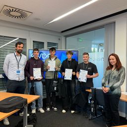 Fünf junge Männer und eine Frau stehen in einem Raum und halten Urkunden in der Hand. Sie lächeln und scheinen eine Errungenschaft zu feiern. Hinter ihnen befindet sich ein Monitor mit Text.