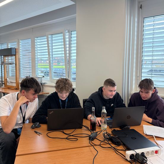 Vier junge Männer sitzen um einen Tisch in einem Büro herum und arbeiten an ihren Laptops. Eine Schreibtischlampe und eine Steckdosenleiste sind auf dem Tisch. Hinter ihnen sind Fenster mit Jalousien.