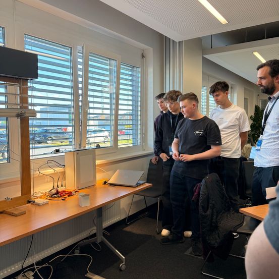 Eine Gruppe junger Männer steht an einem Schreibtisch mit Laptop in einem Raum mit Fenstern und einem Mann mit Ausweiskarte.