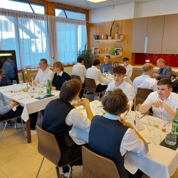Eine Gruppe von Menschen sitzt um einen Tisch in einem Esszimmer. Jede Person hat ein Glas Wasser, eine Kerze und eine Speisekarte vor sich. Der Raum ist mit weißen Vorhängen, Holzschränken und Regalen mit Pflanzen dekoriert.