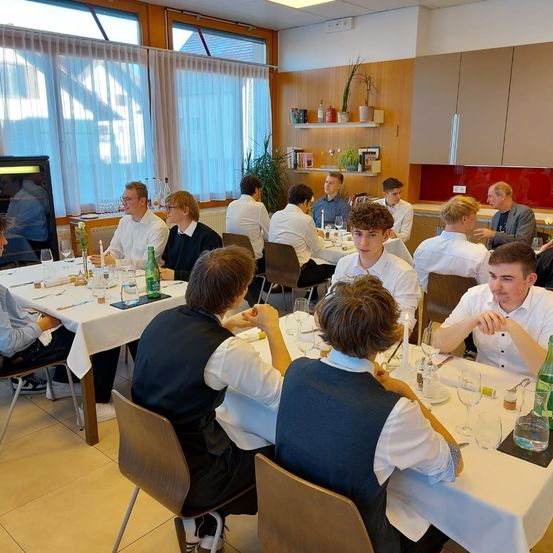 Eine Gruppe von Menschen sitzt um einen Tisch in einem Esszimmer. Jede Person hat ein Glas Wasser, eine Kerze und eine Speisekarte vor sich. Der Raum ist mit weißen Vorhängen, Holzschränken und Regalen mit Pflanzen dekoriert.
