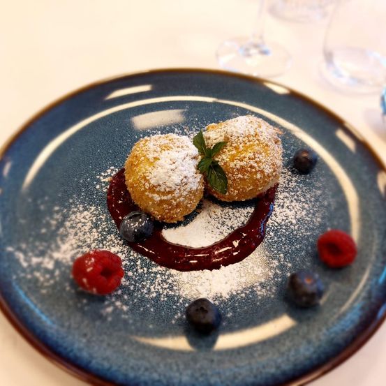 Zwei goldene Donuts mit Puderzucker bestreut, mit roter Beerensauce, Minze, Himbeeren und Blaubeeren garniert, auf einem blauen Teller angeordnet.