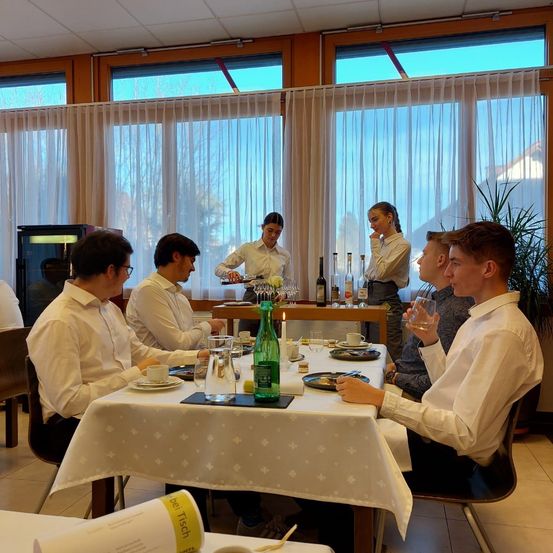 Eine Gruppe von Menschen sitzt an einem Tisch in einem Restaurant. Eine Frau steht hinter ihnen und serviert Wein. Der Tisch ist mit Tellern, Tassen und Weinflaschen gedeckt. Im Hintergrund befinden sich Pflanzen und ein Kühlschrank.