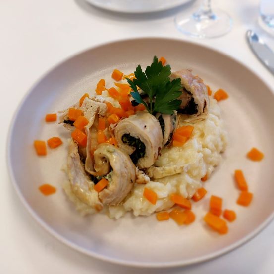 Ein Teller mit cremiger Polenta, geschnittenem Hähnchen, garniert mit gehackten Karotten und einem Petersilienblatt, serviert auf einem weißen Tisch mit Besteck.