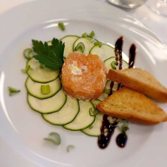 Auf einem Teller befindet sich ein Lachsroulade, umgeben von Gurkenscheiben, garniert mit grünen Kräutern und mit einer dunklen Sauce beträufelt. Zwei Scheiben Toastbrot liegen nebenan.