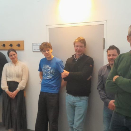 Fünf Personen stehen in einem Raum. Eine Frau in einem weißen Pullover und ein Mann in einem blauen T-Shirt stehen nebeneinander. Ein Mann in einem schwarzen Pullover hat die Arme verschränkt. Hinter ihnen ist eine Tür mit einer Lampe oben.