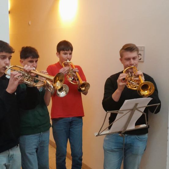 Vier junge Männer spielen Trompeten in einem Flur. Einer liest Noten. Sie sind lässig gekleidet.