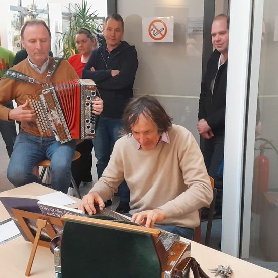 Ein Mann spielt Akkordeon, während ein anderer Mann ihn vom Schreibtisch aus beobachtet. Hinter ihnen stehen drei Personen. Die Umgebung umfasst ein Rauchverbotsschild und eine Glastür.