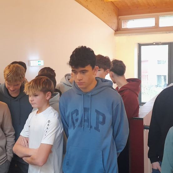 Mehrere junge Jungen stehen in einem Raum, einige tragen Kapuzenpullover. Ein Junge mit einem blauen Hoodie, auf dem 'GAP' steht, sticht hervor. Sie scheinen einem Sprecher zuzuhören.