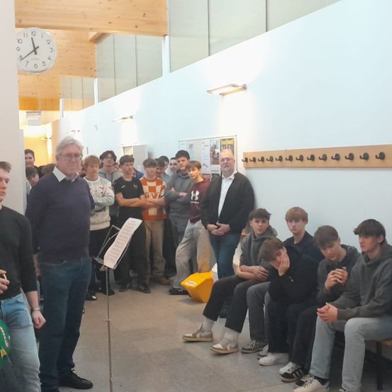 Eine Gruppe von Menschen in einem Flur, einer steht mit einem Notenständer, andere sitzen auf einer Bank. Eine Uhr und Kleiderhaken sind an der Wand.