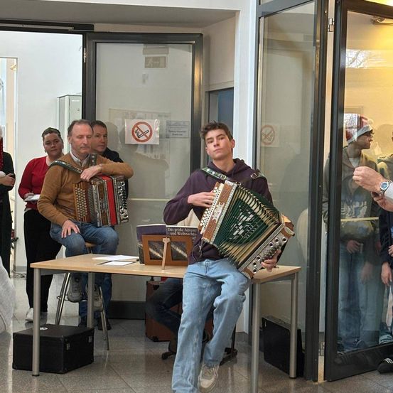 Zwei Männer spielen Akkordeons in einem Raum mit Glaswänden. Ein Mann sitzt auf einem Stuhl, während der andere steht, und mehrere Leute beobachten.