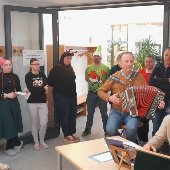 Eine Gruppe von Menschen ist drinnen versammelt. Ein Mann in der Mitte spielt Akkordeon. Eine Frau steht in der Nähe des Eingangs und hält ein Papier. Ein Mann hinter ihr trägt einen Weihnachtsmannhut. Es stehen mehrere Menschen um sie herum.