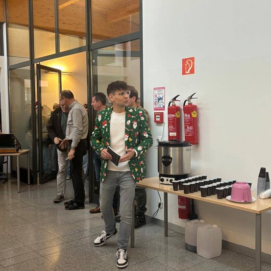 Ein Mann in einem Weihnachtspullover geht an einem Feuerlöscher vorbei. Andere warten in der Schlange. Ein Tisch hält Tassen, einen Kessel und Flaschen. Glastüren und eine Deckenbeleuchtung sind sichtbar.