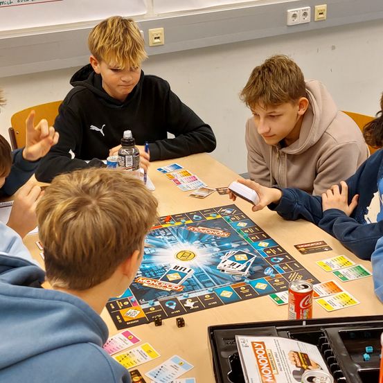 Eine Gruppe von Jungen sitzt um einen Tisch und spielt ein Brettspiel. Das Spiel ist Monopoly, und ein Junge hält einen Würfel.