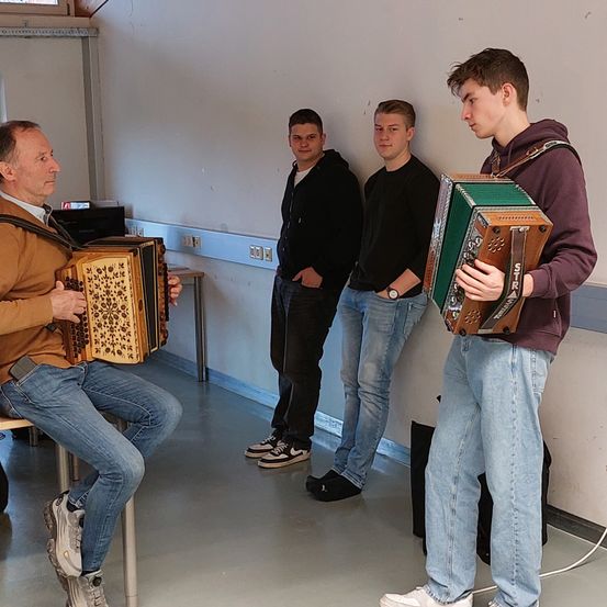 Ein älterer Mann spielt Akkordeon, während drei junge Männer hinter ihm stehen, einer hält ein Akkordeon. Sie sind in einem Raum mit weißer Wand, einem Tisch und Steckdosen.