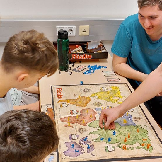 Drei Jungen spielen das Brettspiel Risko. Zwei von ihnen sitzen am Tisch, während der dritte sich über das Brett beugt.