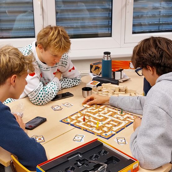 Drei Jungen sitzen um einen Tisch herum und spielen ein Labyrinth-Brettspiel. Ein Junge trägt eine Brille und hält ein Spielstein, ein anderer Junge beobachtet ihn, und ein dritter Junge hält ein Handy. Der Tisch hat ein Labyrinthbrett, Becher und einen Thermos. Das Fenster hinter ihnen hat Jalousien.