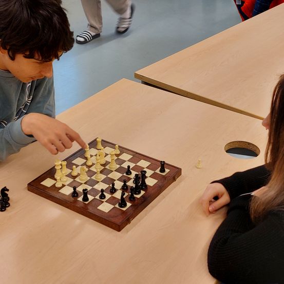 Ein Junge und ein Mädchen spielen Schach in einem Raum mit grauen Böden und Holztischen. Der Junge zeigt auf ein Stück auf dem Schachbrett.