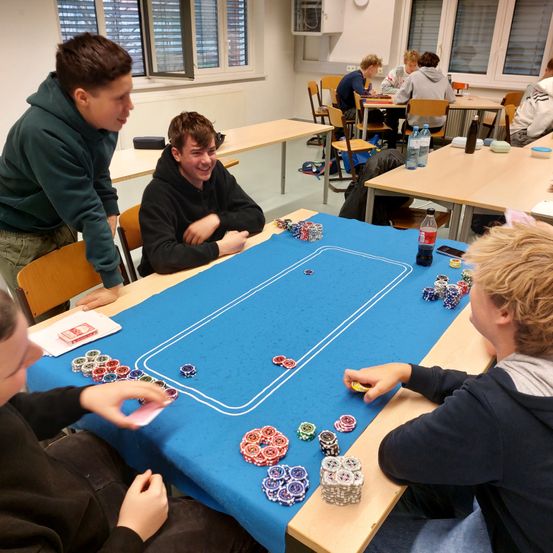 Vier Jungen sitzen um einen Pokertisch mit blauem Tuch. Ein Junge steht, lächelt, während ein anderer Junge mit verschränkten Armen sitzt. Der Tisch hat Pokerchips und Karten.